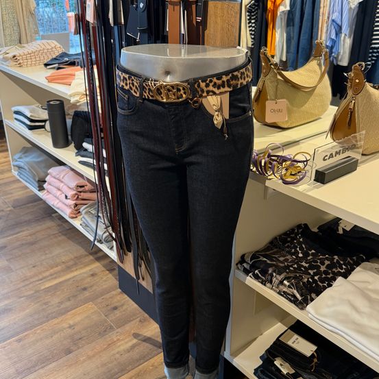 Ein Mannequin in einem Bekleidungsgeschäft zeigt ein Paar dunkle Jeans mit einem Leopardenmustergürtel. Dahinter befinden sich Regale mit verschiedenen Artikeln, darunter Handtaschen, Gürtel und Kleidung.