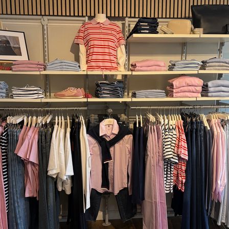 Ein Bekleidungsgeschäft zeigt gefaltete Kleidung auf Regalen und einem Mannequin mit einem rot gestreiften Hemd. Verschiedene farbige Kleidungsstücke hängen an Kleiderbügeln.