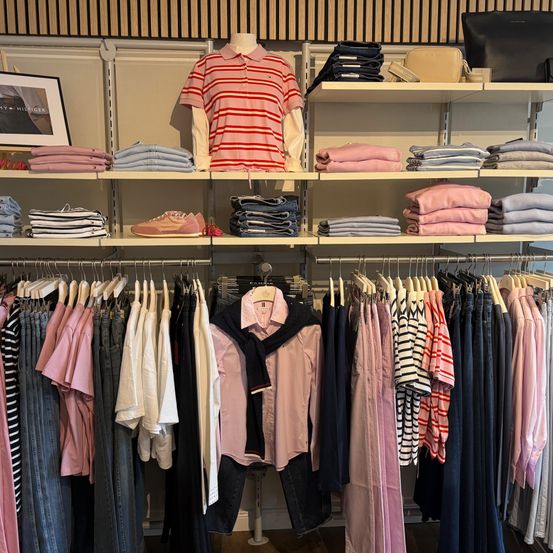 Ein Bekleidungsgeschäft zeigt gefaltete Kleidung auf Regalen und einem Mannequin mit einem rot gestreiften Hemd. Verschiedene farbige Kleidungsstücke hängen an Kleiderbügeln.