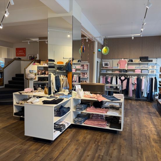 Ein Bekleidungsgeschäft mit Holzboden, weißen Regalen und Kleidung an Kleiderbügeln. Eine Treppe führt in den oberen Stock und ein rotes Schild mit dem Wort Verkauf ist sichtbar.