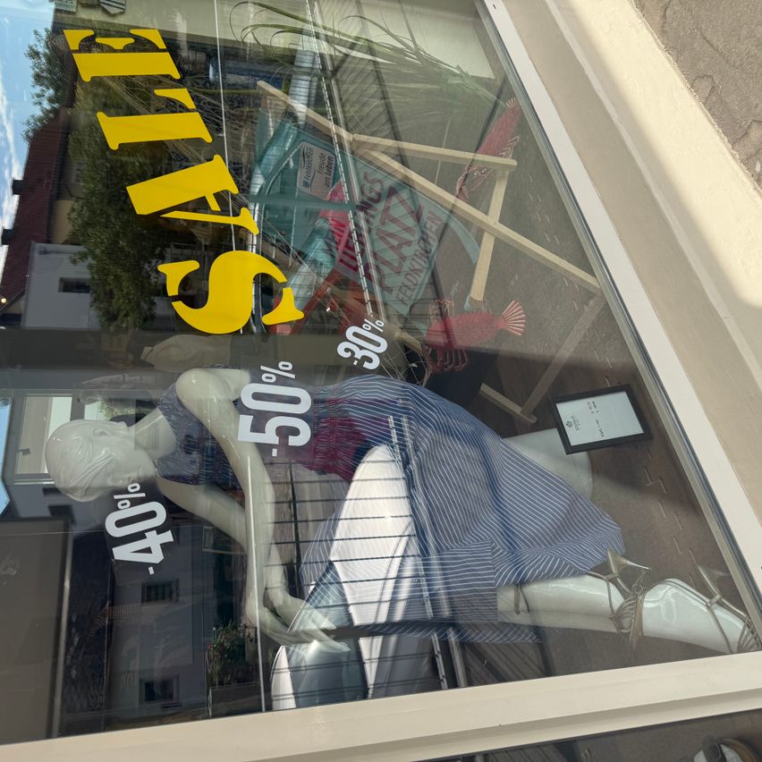 A storefront window displays a mannequin in a blue dress with a price tag reading '40% off'. The window also features a yellow sign with the word 'SALE' and a price reduction of 30%. The background shows a reflection of a building and a tree.