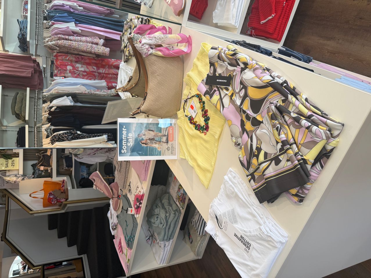 A clothing store displays various summer items, including a yellow shirt, a patterned scarf, and a handbag, all neatly arranged on a white shelf.