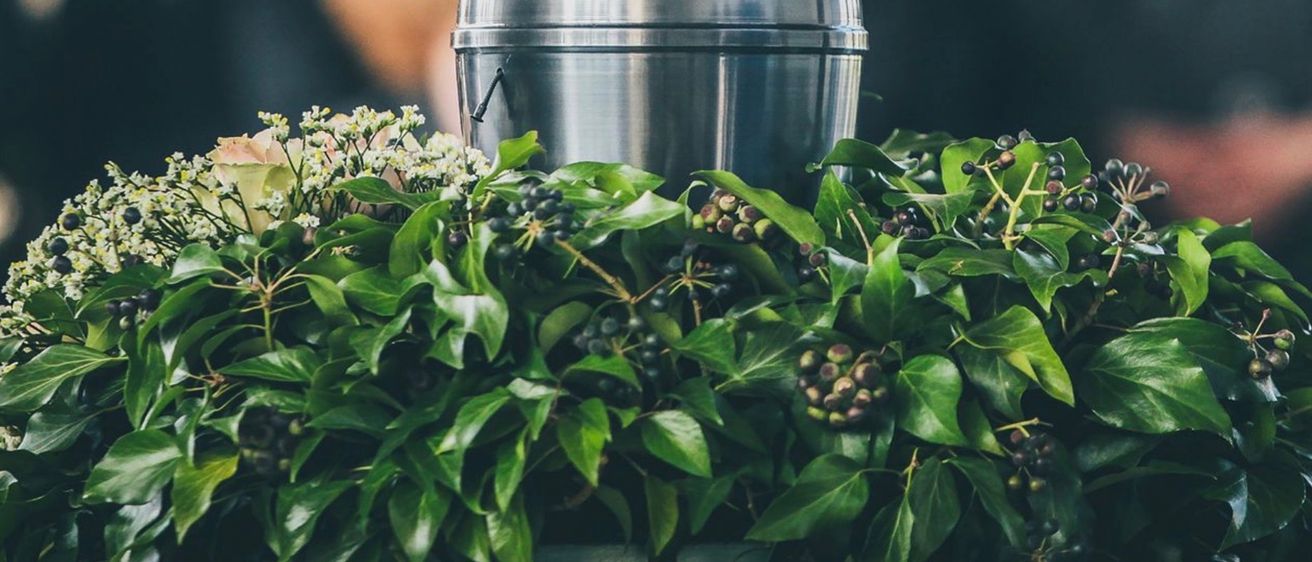 Ein Edelstahlbehälter mit Griff ist teilweise von grünen Blättern und kleinen Beeren bedeckt, mit einer weißen Blume im Hintergrund.