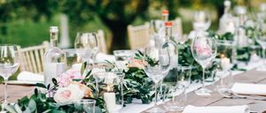 Ein Tisch ist für eine Gartenparty mit Weingläsern, Kerzen und grünen Blättern gedeckt. Flaschen mit roten Deckeln sind vorhanden, und die Szene spielt im Freien.