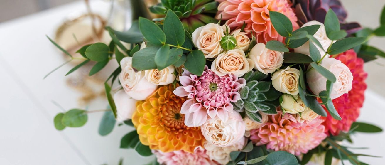 Ein Nahaufnahme eines Hochzeitsbouquets mit einer Mischung aus weißen, cremefarbenen, pfirsichfarbenen und orangefarbenen Rosen, rosa Dahlien und grünem Laub.