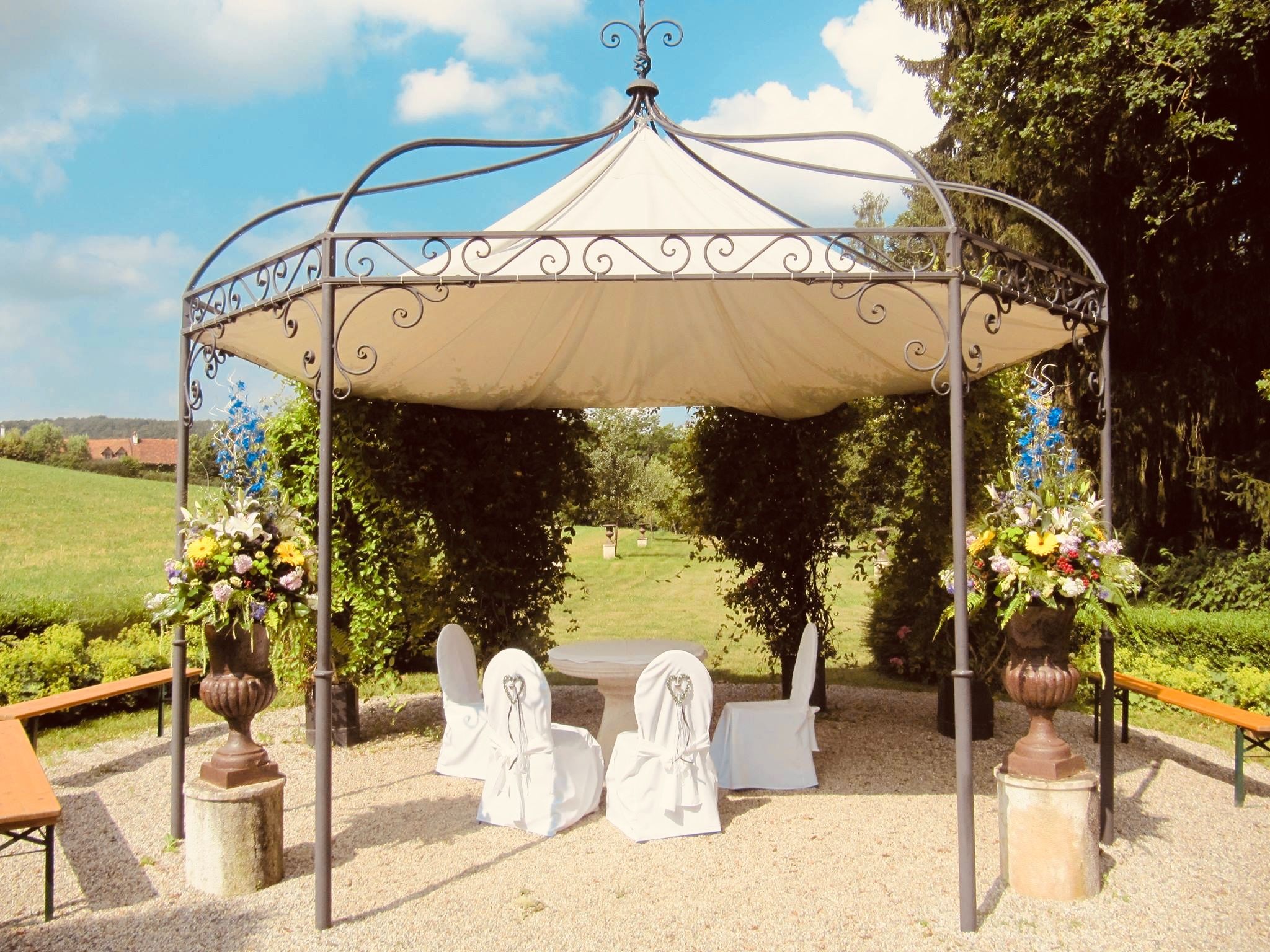 Ein elegantes Metall-Gazebo mit weißem Dach und Blumenvasen steht in einem Garten. Vier Stühle mit Bezüge umgeben einen kleinen Tisch. Der Bereich ist von Grün umgeben.