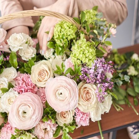 Eine Person, die einen Strauß aus rosa, weißen und lila Blumen in einem Korb auf einem Holztisch arrangiert.