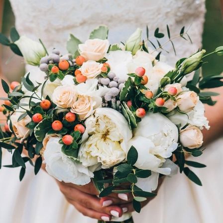 Eine Braut hält einen Hochzeitsstrauß mit weißen und cremefarbenen Blumen, ergänzt durch rote Beeren und grüne Blätter. Das Arrangement wird mit lackierten Nägeln gehalten.