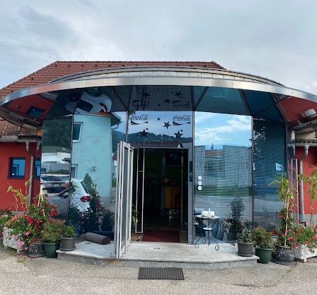 Ein moderner Eingang mit einer Glasüberdachung und Coca-Cola-Markierung. Der Eingang führt zu einer offenen Tür mit einem roten Teppich. Draußen gibt es Blumentöpfe und Pflanzen, mit einem geparkten Auto in der Nähe.