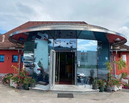 Ein moderner Eingang mit einer Glasüberdachung und Coca-Cola-Markierung. Der Eingang führt zu einer offenen Tür mit einem roten Teppich. Draußen gibt es Blumentöpfe und Pflanzen, mit einem geparkten Auto in der Nähe.