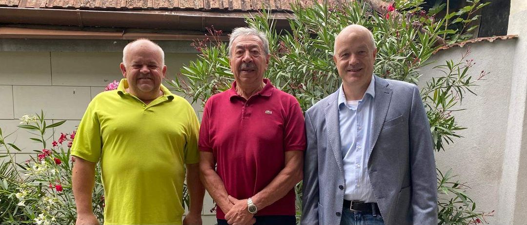 Drei Männer stehen zusammen vor einem Haus mit viel Grün. Einer trägt ein leuchtend gelbes Poloshirt, ein anderer ein rotes Poloshirt und der dritte einen blauen Anzug. Sie alle lächeln in die Kamera.