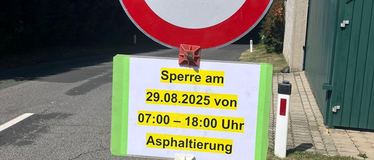 Ein Schild an einem Pfosten mit einem roten Kreis weist auf eine Straßensperrung für Asphaltarbeiten am 29.08.2025 von 07:00 bis 18:00 Uhr hin.