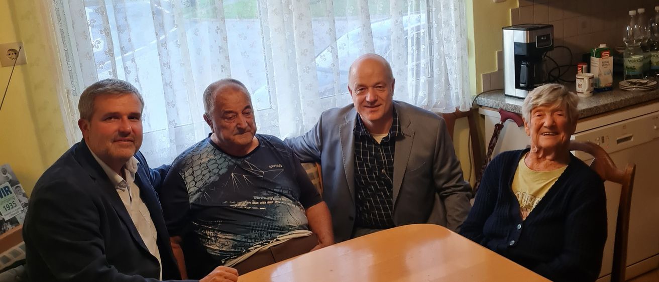 Vier Personen sitzen lächelnd um einen Esstisch herum. Der Mann auf der linken Seite trägt einen dunklen Anzug und Jeans. Der Mann neben ihm trägt ein bedrucktes T-Shirt. Der Mann in der Mitte trägt einen grauen Anzug. Die Frau auf der rechten Seite trägt ein gelbes Hemd und einen Strickpullover. Hinter ihnen befindet sich ein Fenster mit weißen Vorhängen.