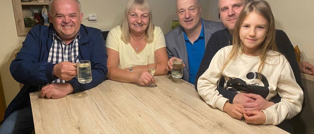 Five people are seated at a table, smiling for a photo. Two men and two women, and a girl, are holding glasses of water. Behind them is a wall with picture frames.
