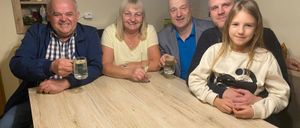 Fünf Menschen sitzen lächelnd an einem Tisch und posieren für ein Foto. Zwei Männer, zwei Frauen und ein Mädchen halten Gläser mit Wasser in der Hand. Hinter ihnen ist eine Wand mit Bilderrahmen.