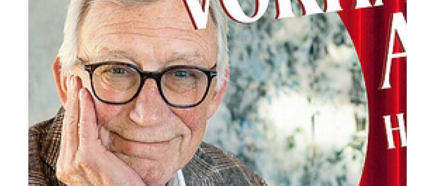 Ein älterer Mann mit Brille lächelt und posiert für ein Foto vor einem roten Vorhang.