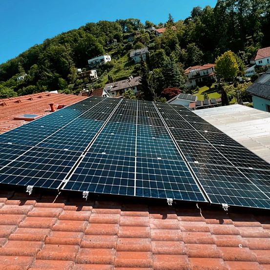 Solarmodule auf einem Dach mit klarem blauem Himmel und Bäumen im Hintergrund. Die Module sind angeschlossen und auf dem Dach installiert.