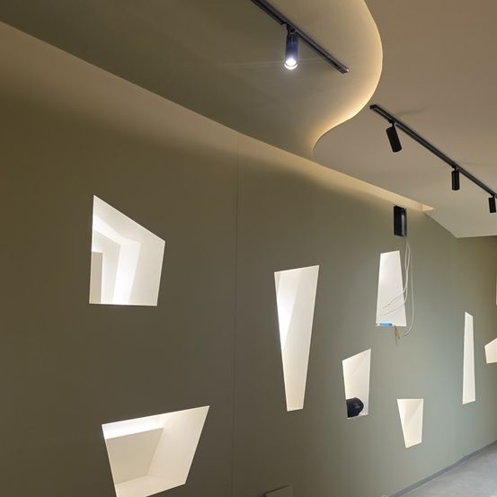 Eine Innenwand mit geometrischen Beleuchtungskörpern; dreieckige Öffnungen lassen Licht ein; die Decke hat eine geschwungene Form mit beleuchteten Schienen.