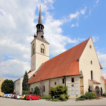 Eine große Kirche mit rotem Dach und einem hohen Turm steht vor einem bewölkten Himmel. Viele Autos sind vor der Kirche geparkt.