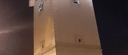 Ein hoher Uhrenturm ist nachts beleuchtet und zeigt zwei Uhren und zwei Sterne. Ein Auto ist am Straßenrand geparkt.