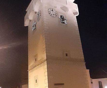 Ein hoher Uhrenturm ist nachts beleuchtet und zeigt zwei Uhren und zwei Sterne. Ein Auto ist am Straßenrand geparkt.