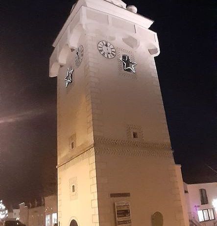 Ein hoher Uhrenturm ist nachts beleuchtet und zeigt zwei Uhren und zwei Sterne. Ein Auto ist am Straßenrand geparkt.
