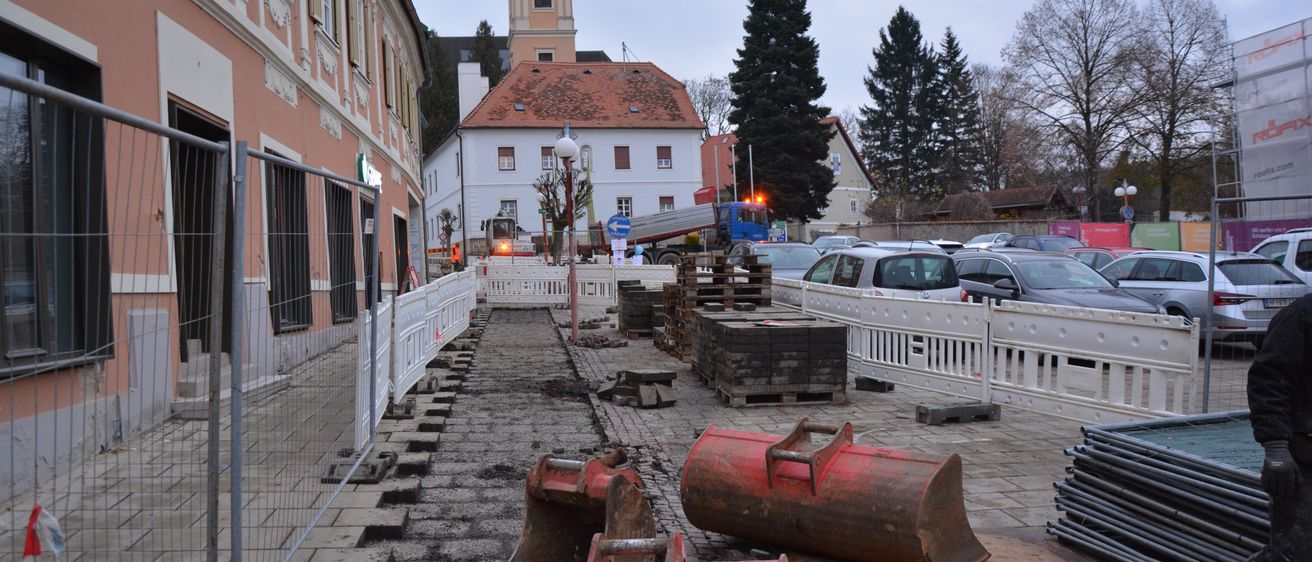 Ein laufendes Baustelle mit einem Gebäude, einem Glockenturm und Autos im Hintergrund. Verschiedene Baumaschinen, einschließlich Bagger, liegen auf dem Boden.