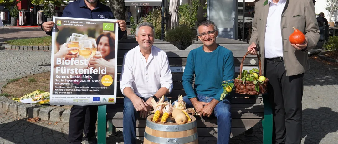 Vier Männer sitzen auf einer Bank, einer hält ein Plakat, ein anderer einen Korb mit Früchten, und zwei weitere lächeln. Sie sind von einem Fass Mais und einem Baum umgeben.