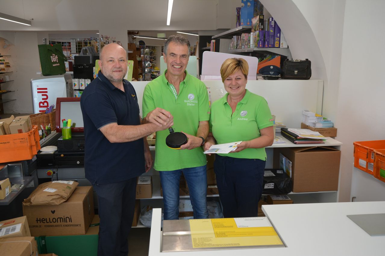 Three people in green shirts stand in a store, with one man holding a black object and smiling. Behind them, shelves hold various items, and a woman holds a piece of paper.