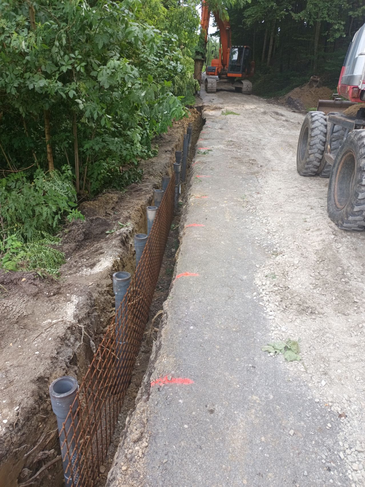 Eine Baustelle mit einer Grube, rostigen Metallzäunen und großen Maschinen. Bäume und Büsche sind sichtbar und der Boden ist uneben.