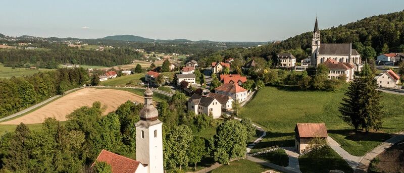Eine Draufsicht auf ein ruhiges Dorf mit Häusern, einer Kirche und grünen Feldern unter einem klaren Himmel.