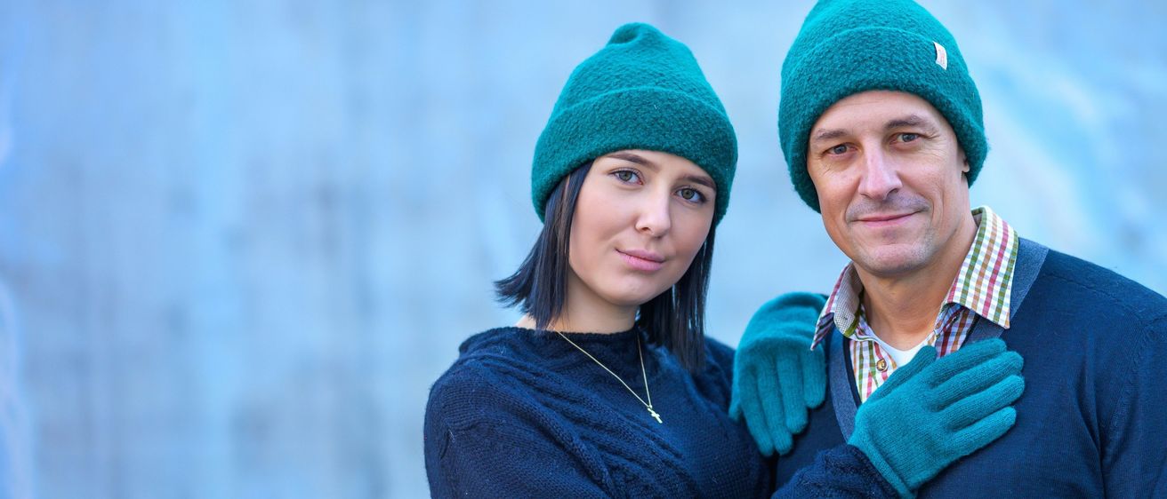 Zwei Personen, ein Mann und eine Frau, stehen zusammen. Sie tragen beide gestrickte türkisfarbene Mützen und lächeln. Die Frau trägt einen blauen Pullover und eine Halskette.