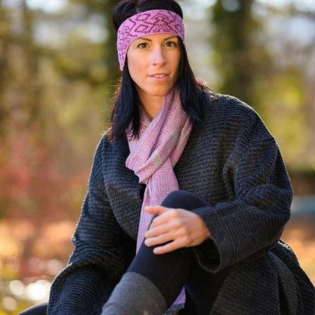 Eine Frau mit einer rosa Kopfband und einem Schal sitzt im Freien, die Hände auf den Knien. Sie trägt einen grauen Pullover und schwarze Leggings. Der Hintergrund ist verschwommen mit Bäumen.