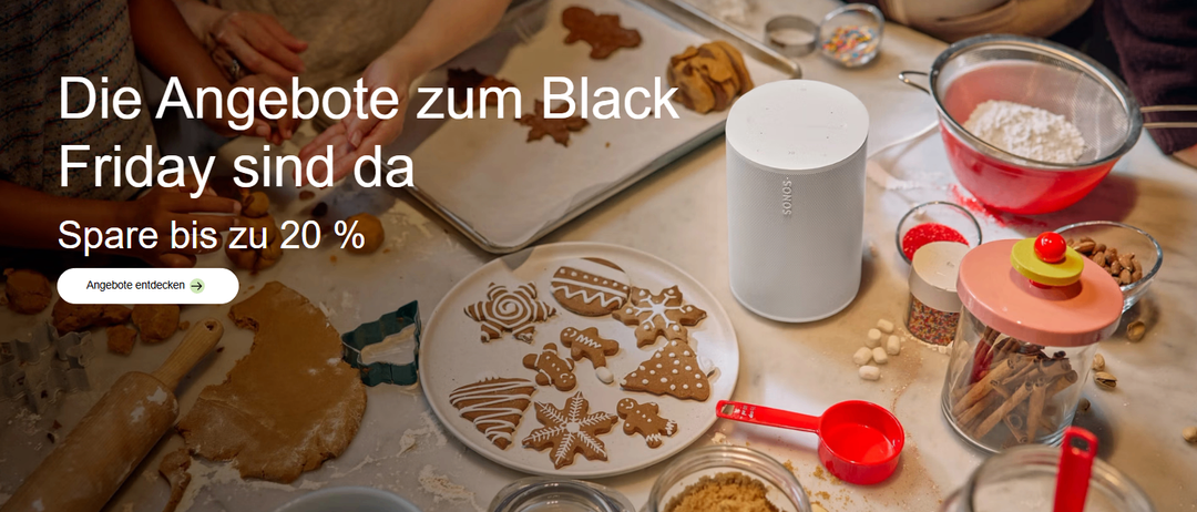 Ein Tisch mit Keksen auf einem Teller und einem Ausstecher. Ein Lautsprecher steht rechts und ein Messbecher links. Eine Hand einer Person liegt auf dem Tisch.