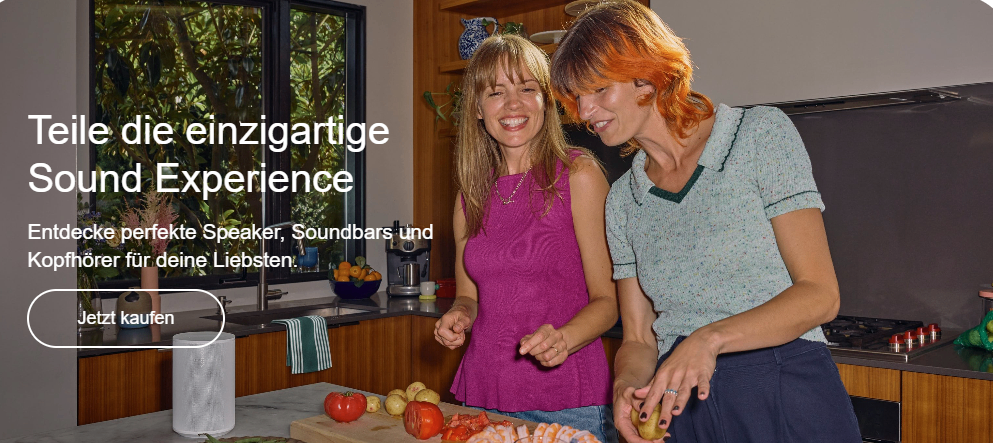 Zwei Frauen in einer Küche lächeln und scheinen Gemüse zu schneiden. Ein Lautsprecher steht auf der Arbeitsplatte und ein Fernseher an der Wand.