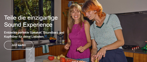 Zwei Frauen in einer Küche lächeln und scheinen Gemüse zu schneiden. Ein Lautsprecher steht auf der Arbeitsplatte und ein Fernseher an der Wand.