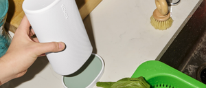 Eine Hand hält einen weißen Sonos-Lautsprecher auf einer Küchentheke. Der Arbeitstisch zeigt ein Holzschneidebrett, Teller, ein Glas und einen grünen Gemüsekorb. Ein Waschbecken mit Wasserhahn und Bürste ist ebenfalls zu sehen.