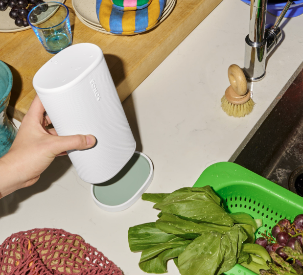 Eine Hand hält einen weißen Sonos-Lautsprecher auf einer Küchentheke. Der Arbeitstisch zeigt ein Holzschneidebrett, Teller, ein Glas und einen grünen Gemüsekorb. Ein Waschbecken mit Wasserhahn und Bürste ist ebenfalls zu sehen.