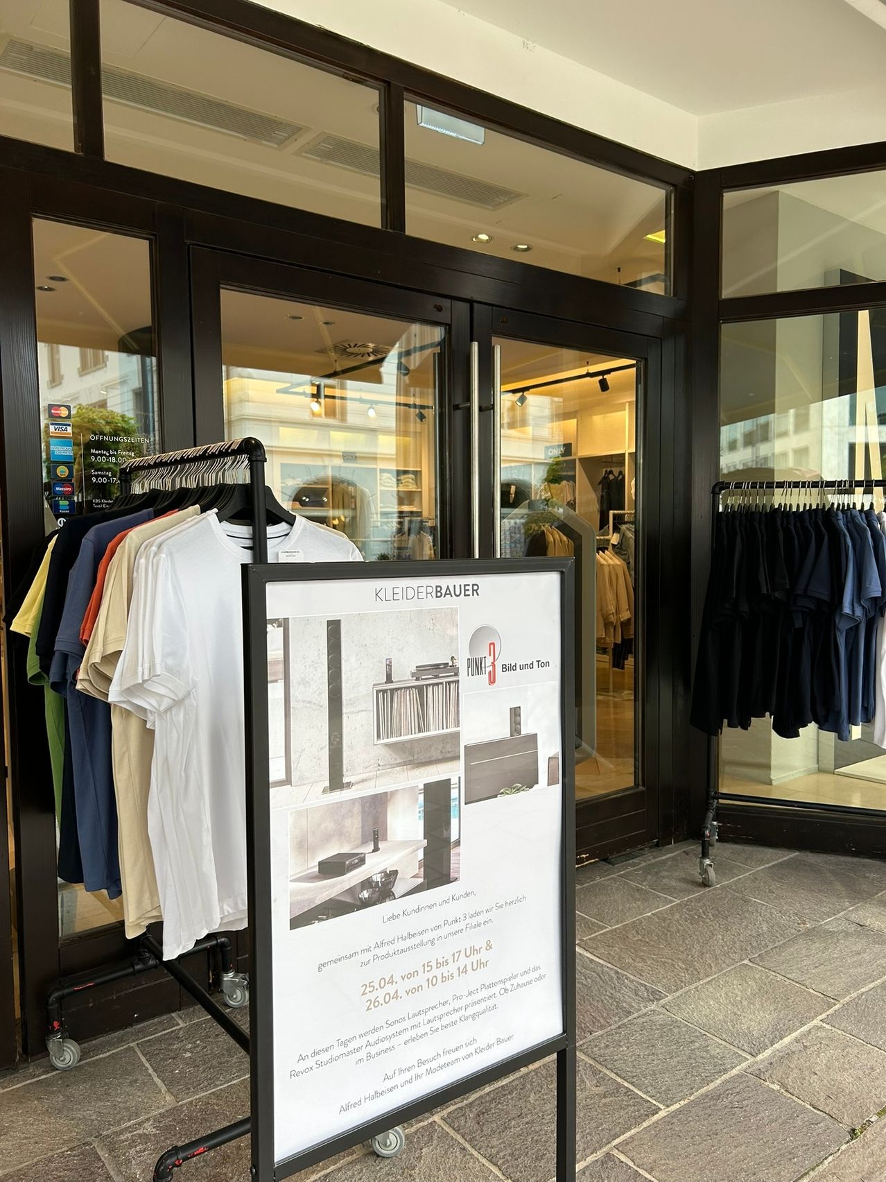 Ein Bekleidungsgeschäft mit einem Ständer für T-Shirts vor dem Eingang und einem Schild mit Zeitplan für den 25. und 26. April.