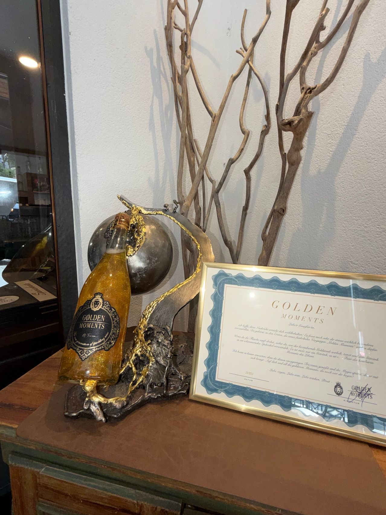 A golden bottle of Golden Moments champagne is displayed on a table next to a certificate. The bottle is placed on a decorative sculpture, and a shadow falls on the wall behind.