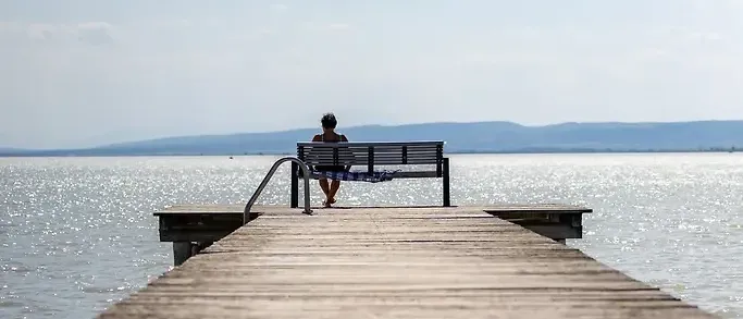 Eine Person sitzt auf einer Bank auf einem hölzernen Steg, mit Blick auf das ruhige Meer und Berge in der Ferne unter einem klaren Himmel.