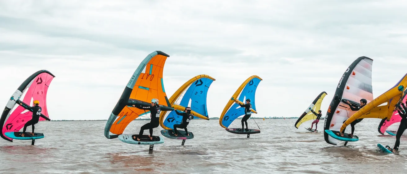Eine Gruppe von Menschen windsurft auf dem Wasser mit bunten Segeln unter einem bewölkten Himmel.