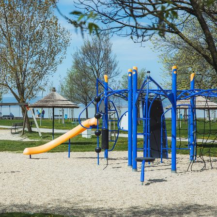 Ein Kinderspielplatz befindet sich auf einer sandigen Oberfläche. Er hat eine Rutsche, Schaukeln und eine Kletterstruktur. Bänke und ein Pavillon befinden sich in der Umgebung. Bäume und Berge sind in der Ferne.