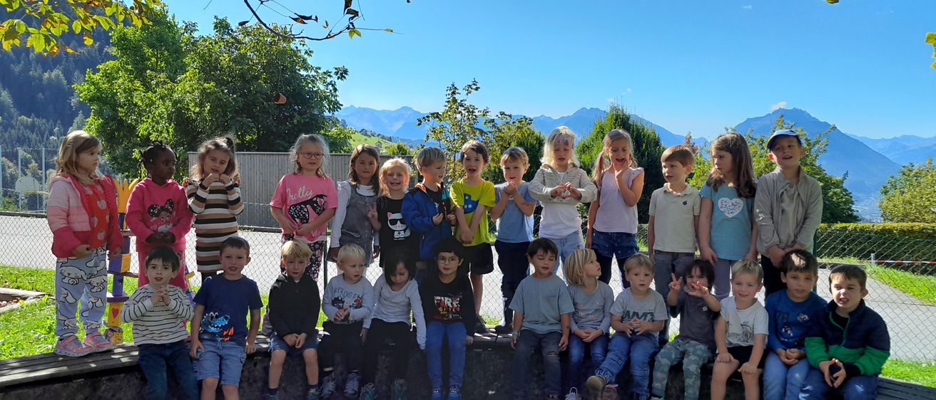 Eine Gruppe von Kindern steht und sitzt auf einer Betonmauer in einem Außenbereich. Sie lächeln und posieren für ein Foto. Hinter ihnen befinden sich Bäume, ein Zaun und Berge.