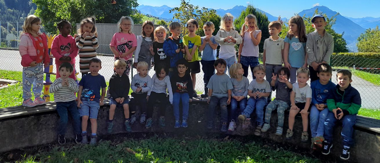 Eine Gruppe von Kindern steht und sitzt auf einer Betonmauer in einem Außenbereich. Sie lächeln und posieren für ein Foto. Hinter ihnen befinden sich Bäume, ein Zaun und Berge.