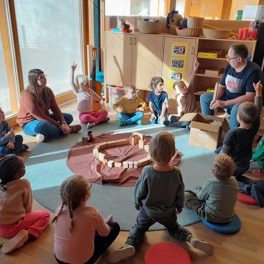 Eine Gruppe von Kindern und zwei Erwachsenen sitzt in einem Kreis um einen Tisch mit Holzklötzen, einer Gitarre und Körben.