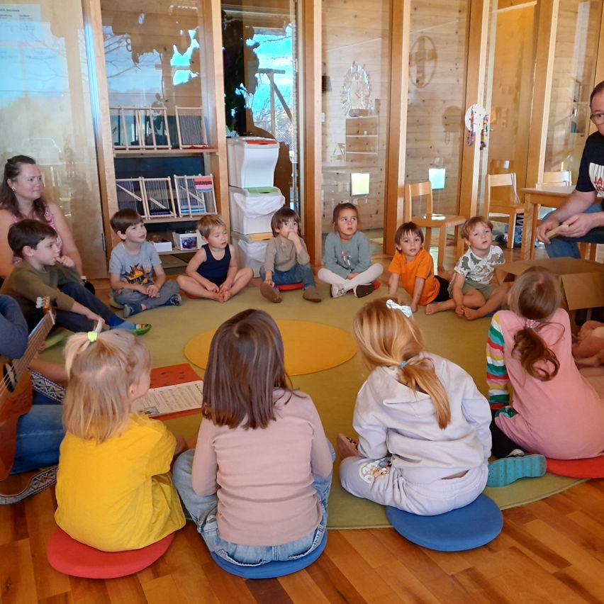 Eine Gruppe von Kindern sitzt im Kreis und hört aufmerksam einer Frau zu, die eine Gitarre spielt. Hinter ihnen ist ein Raum mit Holzwänden und Möbeln zu sehen.