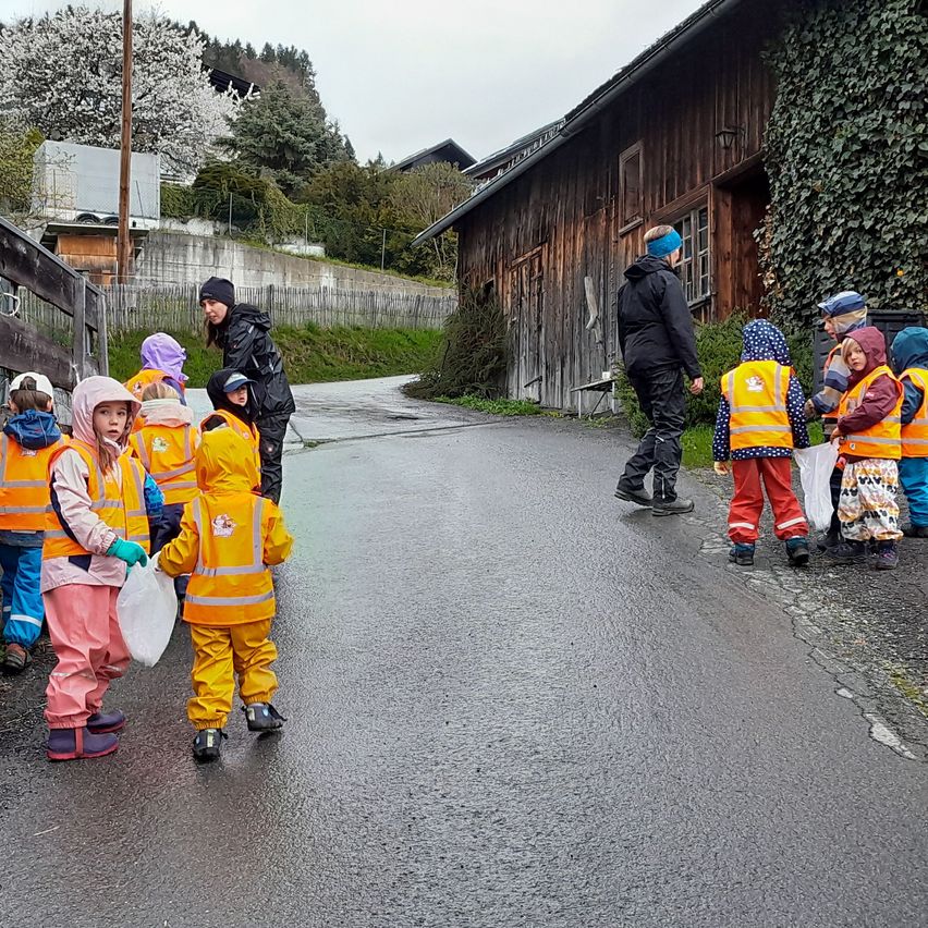 Eine Gruppe von Kindern in orangefarbenen Regenmänteln geht auf einer nassen Straße mit einem Erwachsenen. Im Hintergrund befindet sich ein Holzgebäude und Grünfläche.