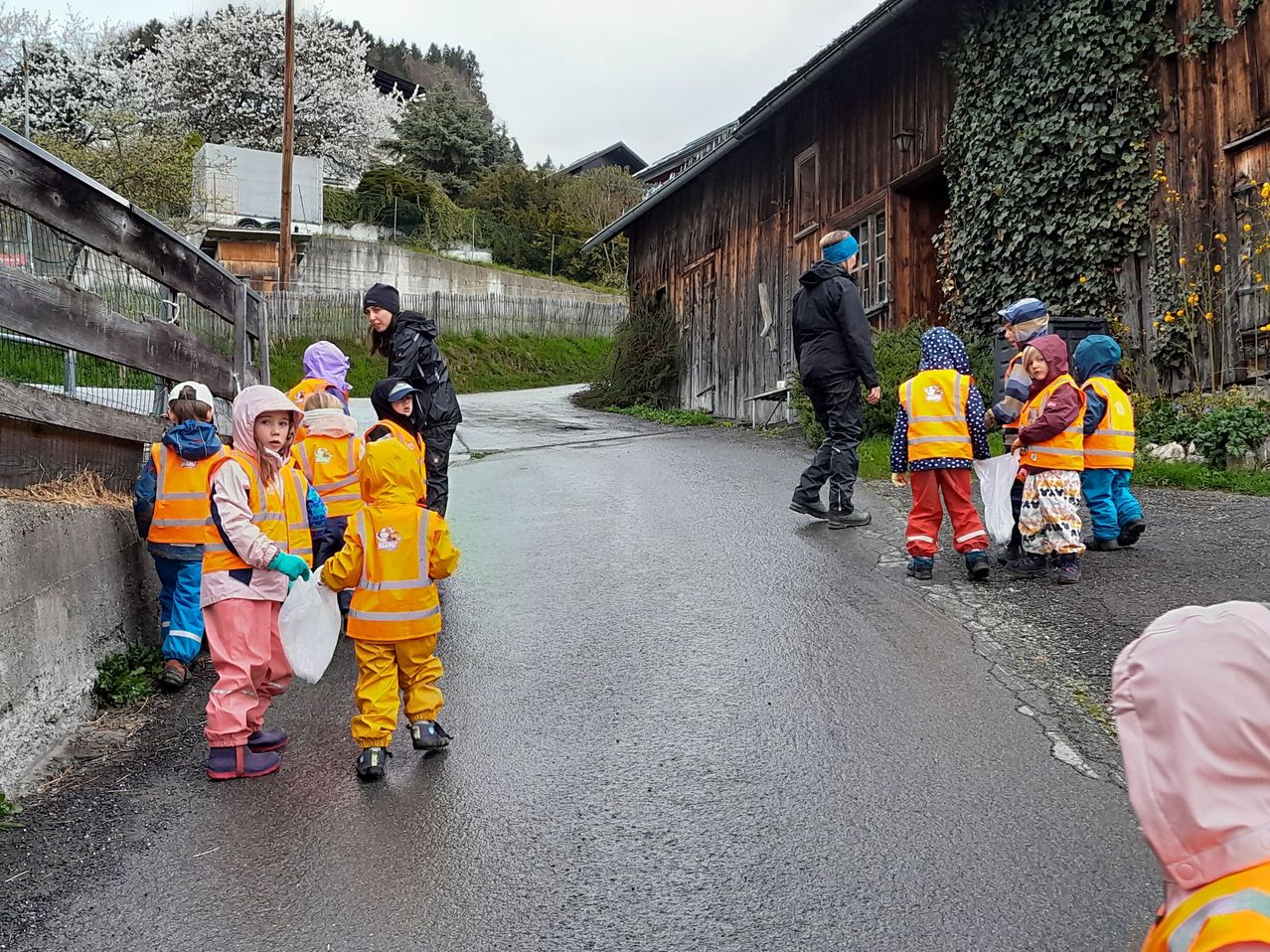 Eine Gruppe von Kindern in orangefarbenen Regenmänteln geht auf einer nassen Straße mit einem Erwachsenen. Im Hintergrund befindet sich ein Holzgebäude und Grünfläche.