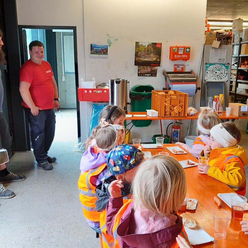 Eine Gruppe von Kindern in Sicherheitswesten isst am Tisch. Ein Mann in einem roten Shirt steht im Hintergrund.